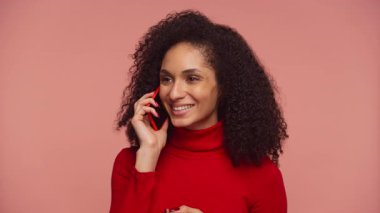 Kırmızı kazaklı neşeli bir kadın akıllı telefonuyla konuşurken gülümsüyor. Pembe arka plandaki bir telefon görüşmesinde hoş bir sohbetin tadını çıkarıyor..