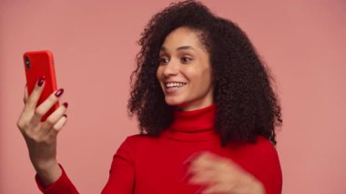 Kırmızı kazaklı neşeli bir kadın, pembe bir arka planda içten bir gülümsemeyle video görüşmesi yaparken akıllı telefonunu tutuyor..