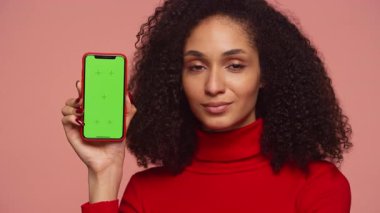 Kırmızı kazaklı neşeli bir kadın yeşil ekranlı bir akıllı telefon tutuyor ve pembe bir arka plana içerik yerleştirmek için kişiselleştirilebilir bir görüntü sunuyor..