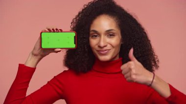 Kırmızı kazaklı neşeli bir kadın yeşil ekranlı bir akıllı telefonu tutuyor ve pembe arka planda kişiselleştirilebilir bir görüntü sunuyor..