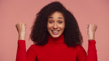 Kırmızı kazaklı neşeli bir kadın heyecan içinde yumruklarını kaldırıyor, başarıyı ve başarıyı pembe arka planda büyük bir gülümsemeyle kutluyor..