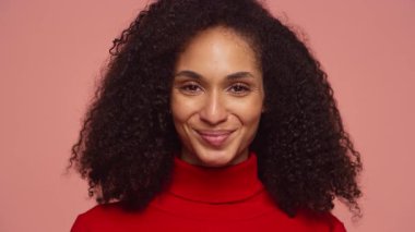 Kıvırcık saçlı, kırmızı balıkçı yaka kazak giyen, yumuşak pembe arka plana sıcak bir şekilde gülümseyen neşeli bir kadının yakın plan portresi..