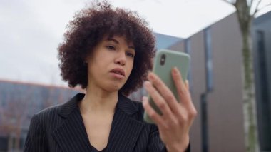 Siyah ceketli genç bir kadın, modern bir ofis binasının önünde telefonuyla video görüşmesi yapıyor. Nişanlı ve ciddi görünüyor..