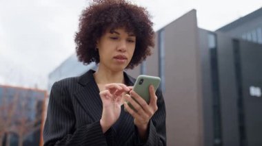 Siyah ceketli genç profesyonel bir kadın modern bir ofis binasının dışındaki akıllı telefonuyla internette geziniyor, odaklanmış ve ciddi görünüyor..