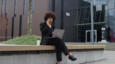 Modern bir ofis binasının önünde laptopuyla konuşurken siyah takım elbiseli odaklanmış profesyonel bir kadın telefonda konuşuyor..