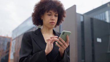 Siyah takım elbiseli profesyonel bir kadın, modern bir ofis binasının önünde dikilirken, akıllı telefonuna odaklanmış, içerikleri karıştırıyor..