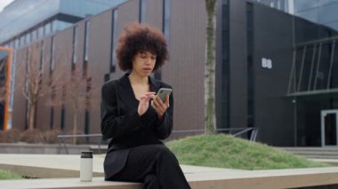 Siyah takım elbiseli neşeli profesyonel bir kadın, modern ofis binasının dışında akıllı telefonuyla gülerken, olumlu içerikten zevk alıyor..