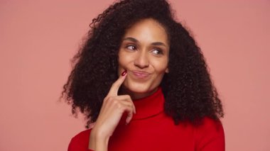 Kırmızı balıkçı yaka yaka giyen genç bir kadın, pembe bir arka plana karşı bir alışveriş yapmayı düşündükçe gülümseyerek parmağını yanağına koyar..