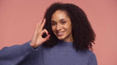 Mavi kazaklı neşeli genç bir kadın pembe bir arka plana el hareketi yaparken ışıl ışıl gülümser, pozitifliğini ve onayını ifade eder..