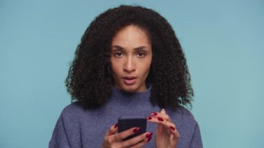 Mavi kazaklı, şaşkın ve şaşkın bakışlı genç bir kadın mavi arka planda beklenmedik haberler okurken akıllı telefonunu tutuyor..