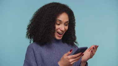 Mavi kazaklı neşeli genç bir kadın, akıllı telefonundan parlak mavi bir arka plana karşı olumlu haberler okurken heyecanla tepki veriyor..