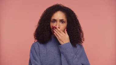 Mavi kazaklı bir kadın eliyle ağzını kapatıyor pembe bir arka plana karşı şok olmuş ve kederli bir ifade sergiliyor..