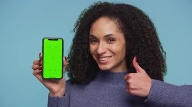 Kıvırcık saçlı neşeli bir kadın yeşil ekranlı akıllı bir telefonu tutarken mavi bir arka planda duruyor..