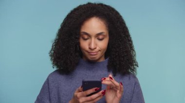 Kıvırcık saçlı bir kadın akıllı telefonuna şüpheci bir ifadeyle bakar, muhtemelen online alışveriş fiyatlarına ya da ürün değerlendirmelerine tepki verir..