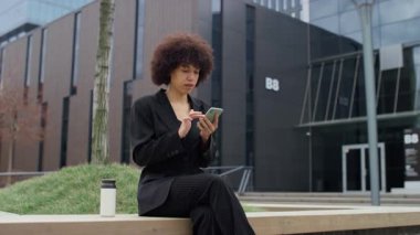 Siyah takım elbiseli profesyonel bir kadın ofis binasının önünde oturuyor. Mola sırasında internetten telefonuna bakıp gülümsüyor..