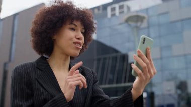 Siyah takım elbiseli profesyonel bir kadın video görüşmesinde modern bir ofis binasının önünde akıllı telefonunu kullanarak el kol hareketi yapar..
