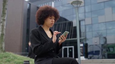 Siyah takım elbiseli profesyonel bir kadın, İnternet 'te çalışırken ya da internette dolaşırken akıllı telefonuna odaklanmış modern bir ofis binasının önünde oturuyor..