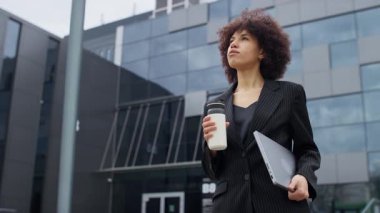 Siyah takım elbiseli profesyonel bir kadın, elinde bir dizüstü bilgisayar ve kahve fincanıyla modern bir ofis binasının önünde yürüyor..