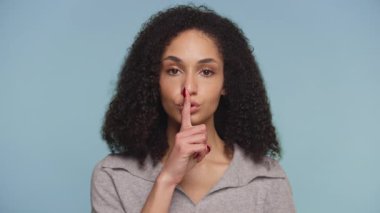 Kıvırcık saçlı genç bir kadın, parmağını dudaklarına dayayıp, mavi arka planda sessizlik ya da gizlilik işareti yapar..