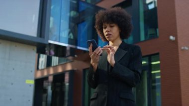 Siyah çizgili takım elbiseli endişeli bir iş kadını, modern bir ofis binasının önünde akıllı telefonundan kötü haberler okurken şok geçirir..