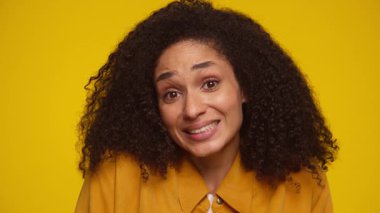 Kıvırcık saçlı, hardal sarısı tişörtlü genç bir kadın tuhaf bir şekilde gülümsüyor, sarı arka planda rahatsızlık veya belirsizlik gösteriyor..