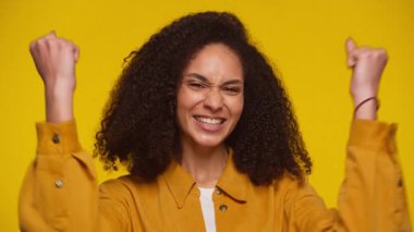 Hardal sarısı tişörtlü, kıvırcık saçlı neşeli bir kadın heyecanlı bir şekilde yumruklarını kaldırıyor ve canlı sarı bir arka plana karşı başarısını kutluyor..