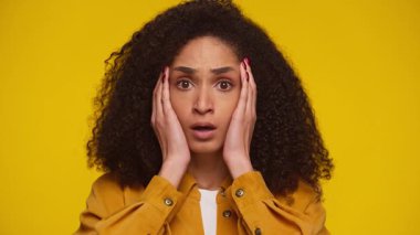 Kıvırcık saçlı, hardal sarısı gömlek giyen genç bir kadın, sarı bir arka plana karşı yüzünü inançsızlıkla tutarak şok olmuş ve şaşırmış görünüyor..