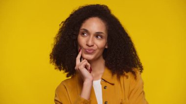 Hardal sarısı tişört giyen kıvırcık saçlı genç bir kadın sarı arka planda şakacı bir ifade takınırken yüzüne dokunuyor..