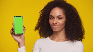Bir kadın yeşil ekranlı bir akıllı telefonu sarı arka planda tutarken kafasını sallıyor..