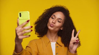 Kıvırcık saçlı, hardal sarısı tişörtlü neşeli bir kadın akıllı telefonuyla selfie çeker barış işareti yapar ve dudaklarını sarı arka plana büzer..