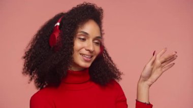 Kıvırcık saçlı, kırmızı kazak giyen ve kulaklık takan neşeli bir kadın müzik eşliğinde dans ediyor, gülümsüyor ve pembe arka plan karşısında anın tadını çıkarıyor..