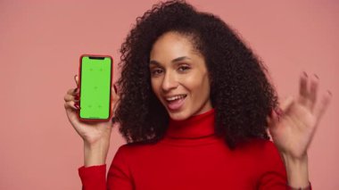 Kırmızı kazaklı neşeli bir kadın yeşil ekranlı bir akıllı telefonu tutuyor ve pembe arka planda kişiselleştirme için boş bir görüntü sergiliyor..
