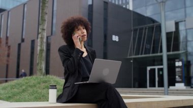 İnce çizgili takım elbiseli genç profesyonel kadın, bilgisayarını modern bir ofis binasının önünde çalışırken telefonla konuşuyor..