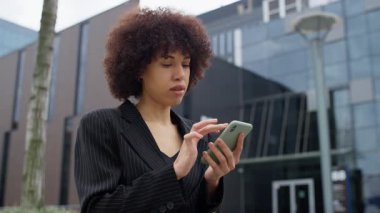 İnce çizgili takım elbiseli genç profesyonel bir kadın modern bir ofisin önünde dururken akıllı telefonuyla içeriği karıştırıyor..