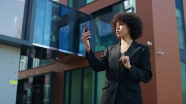 İnce çizgili takım elbiseli genç profesyonel bir kadın modern bir ofis binasının önünde dikilirken akıllı telefonuyla bir video görüşmesi yapıyor..