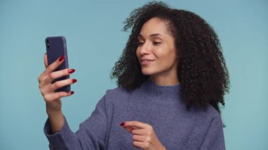 Kıvırcık saçlı, gülümseyen genç bir kadın, akıllı telefonuyla konuşurken mavi bir arka planda..