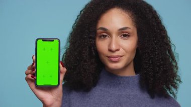 Kıvırcık saçlı, gülümseyen ve onaylayan genç bir kadın mavi arka planda yeşil ekranlı bir akıllı telefonu tutarken başını sallıyor..