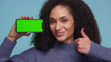 Kıvırcık saçlı, gülümseyen bir kadın, mavi arka planda yüzünün yanında yeşil ekranlı bir akıllı telefonu tutarken baş parmağını kaldırır..