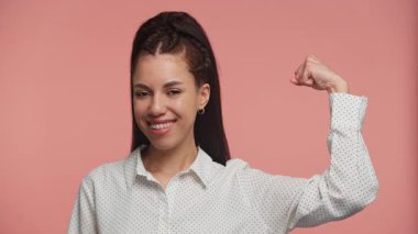 Güçlü genç bir kadın gururla gülümserken güçlü bir poz veriyor, diş telleri takıyor ve pembe arka planda beyaz bir gömlek giyiyor..