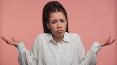 Şaşkın bakışlı genç bir kadın omuzlarını silkiyor ve ellerini kaldırıyor. Pembe bir arka planda kafa karışıklığı ya da şüphe ifade ediyor..