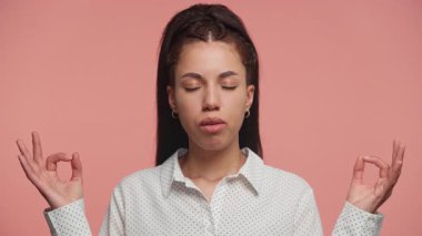 Huzurlu genç bir kadın gözleri kapalı meditasyon yapıyor ve elleri bir Mudra jestinde, pembe arka planda rahatlama ve farkındalık gösteriyor..