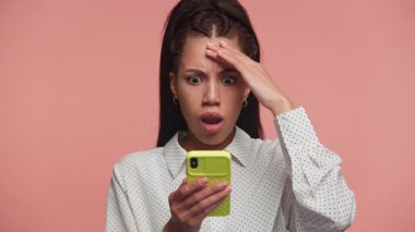 Akıllı telefonuna geniş gözlü ve alnına bakan genç bir kadın beklenmedik haberlere tepki veriyor, pembe arka plana karşı..