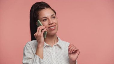 Gülümseyen genç bir kadın, hoş bir telefon konuşması yapıyor, kulağına bir akıllı telefon dayıyor, yumuşak pembe bir arka plana karşı..