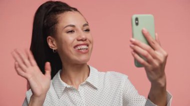 Video görüşmesi için akıllı telefonu kullanan neşeli genç bir kadın. Pembe arka planda sohbet ederken gülümsüyor ve el sallıyor..