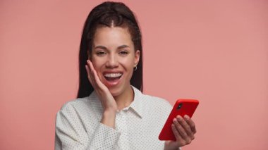 Kırmızı bir akıllı telefon tutan neşeli genç bir kadın sürpriz ve mutlulukla tepki veriyor. Yüzü açık gülümsüyor, pembe arka planda soyutlanıyor..