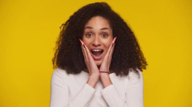 Heyecanlı genç bir kadın. Elleri yanaklarında şok ya da şaşkınlık içinde. Fotokopi çekilen parlak sarı arka plandaki kameraya bakıyor..