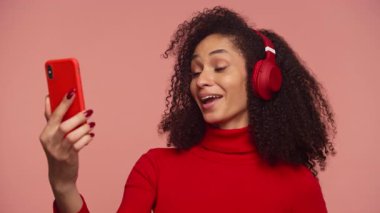 Kulaklık takan ve video görüşmesi sırasında konuşan, elinde akıllı telefon tutan ve pembe arka planda fotokopi çeken genç bir kadın..