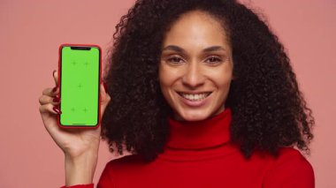 Elinde yeşil ekran ekranı olan akıllı telefonu tutan mutlu genç bir kadın. Pembe arka planda fotokopi alanı olan kameraya gülümsüyor..