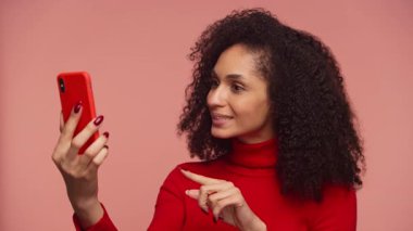 Genç bir kadın, elinde akıllı bir telefonla bir video görüşmesi yapıyor ve pembe arka planda fotokopi çekilen anlamlı jestlerle konuşuyor..