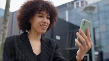 Video görüşmesi sırasında gülümseyen ve elinde akıllı telefonuyla çağdaş bir ofis binasının önünde duran genç profesyonel bir kadın..